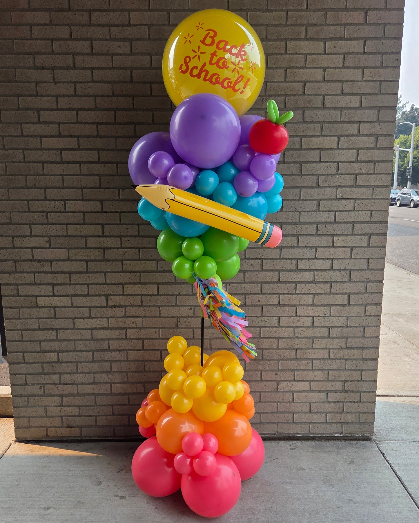 A colorful balloon decoration stand featuring a 'Back to School' yellow balloon, purple and green balloons, a yellow pencil, a red apple, and orange and pink balloons at the base, set against a brick wall.